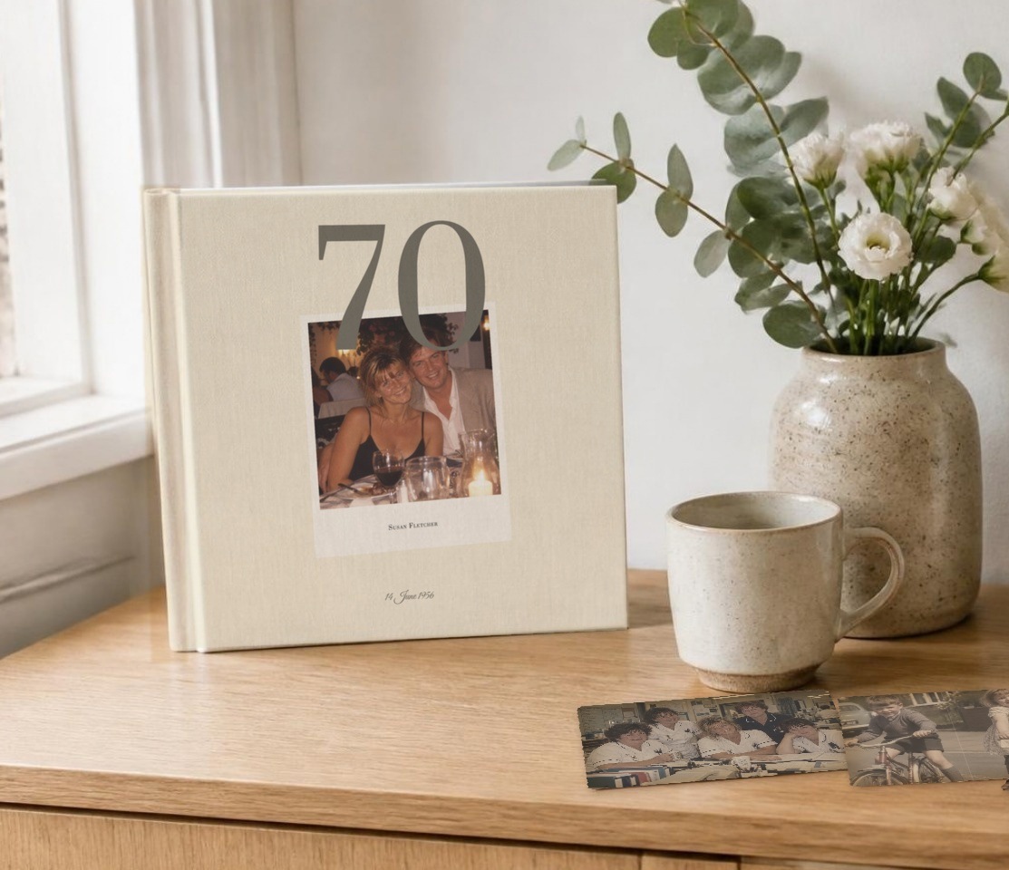 A finished birthday book standing on a sideboard beside a vase of flowers and old photographs.